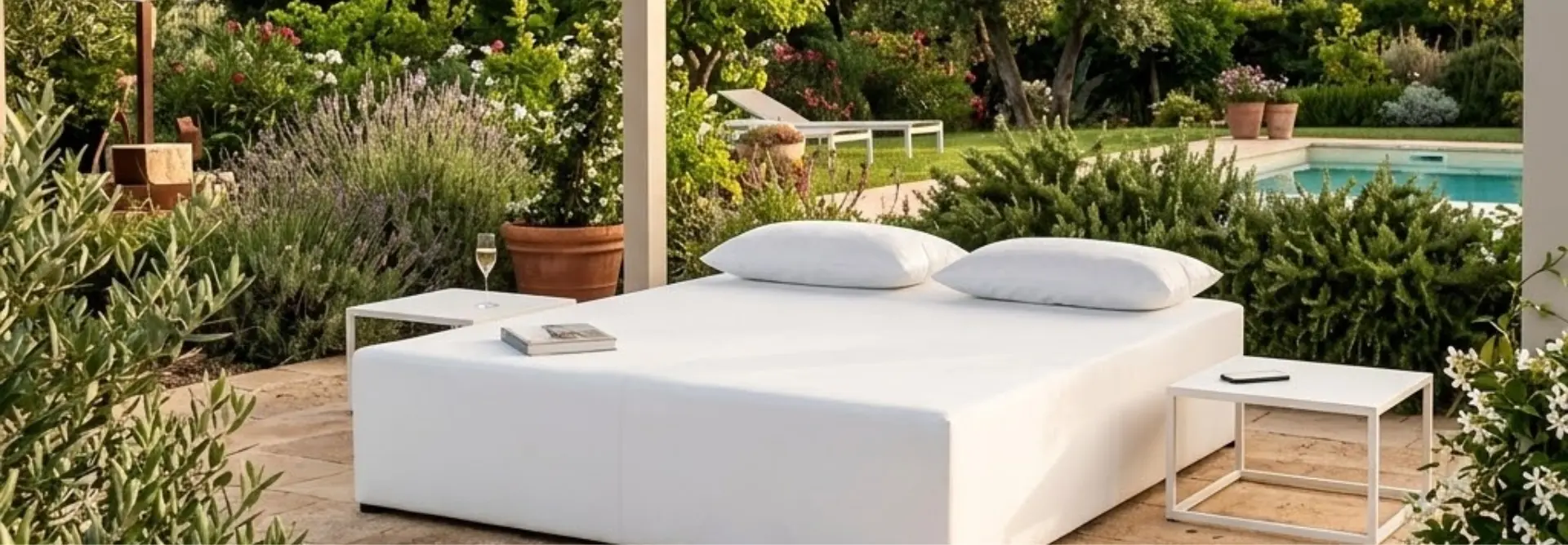 Grande daybed bianco da esterno con cuscini su un patio in pietra, circondato da un giardino mediterraneo con piscina sullo sfondo.